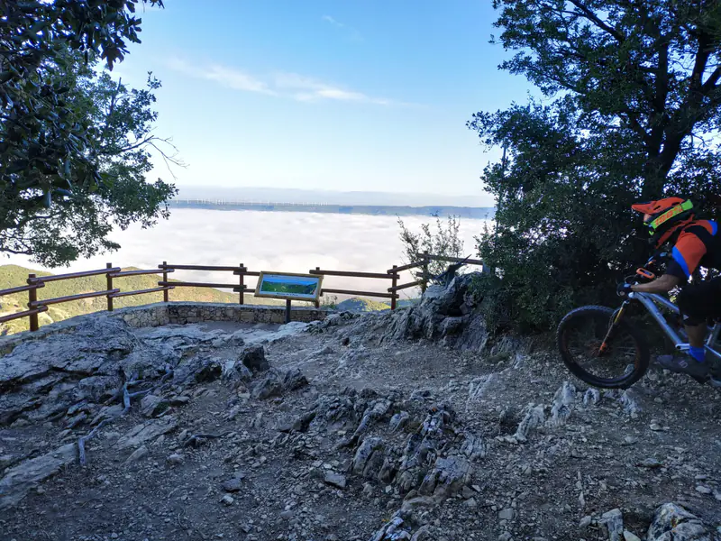 Ger riding an enduro bike into a rocky descent at a viewpoint, above a sea of clouds